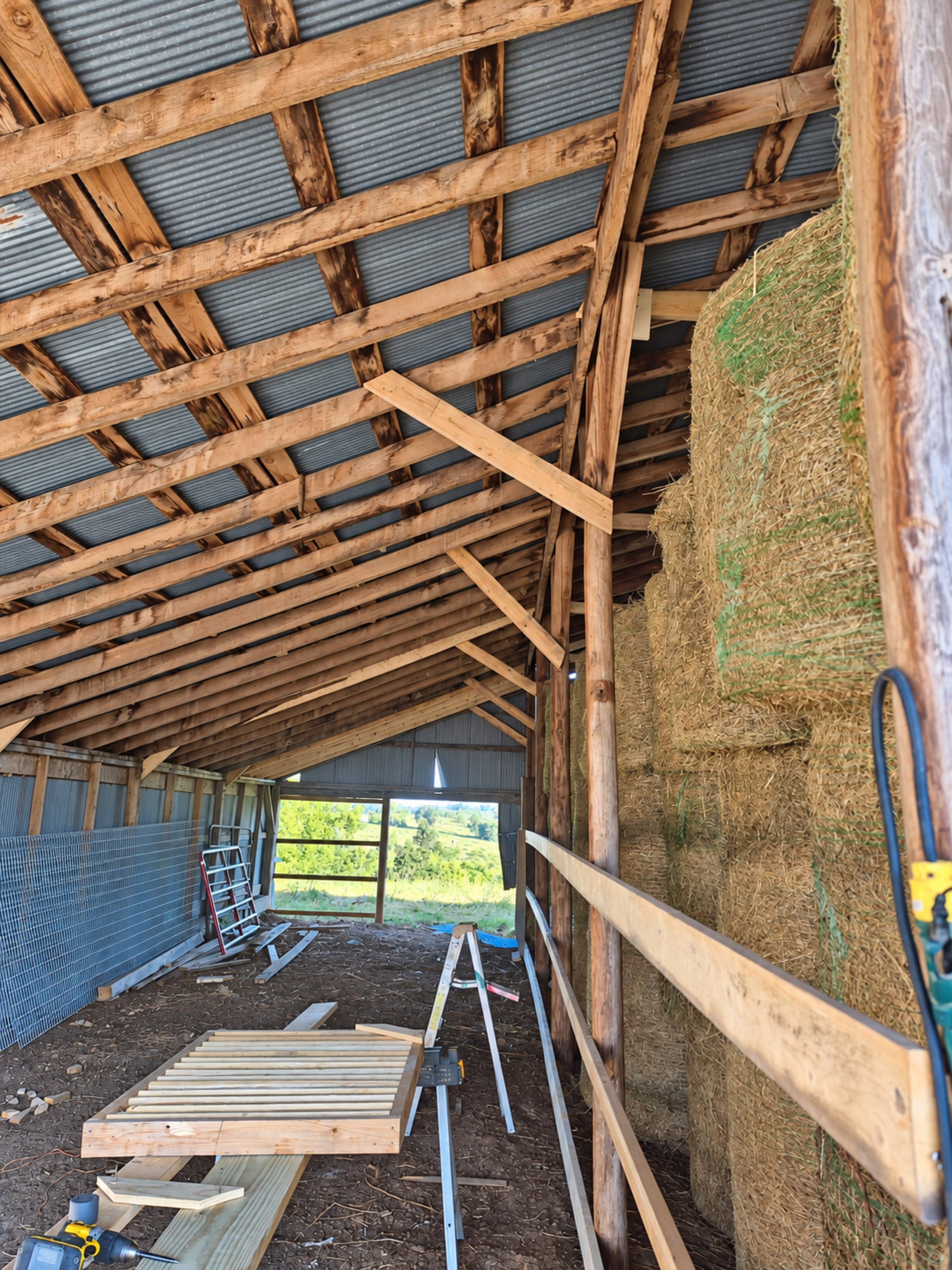 Barn aisle beside stacked hay bales with timber roof framing and fresh diagonal braces